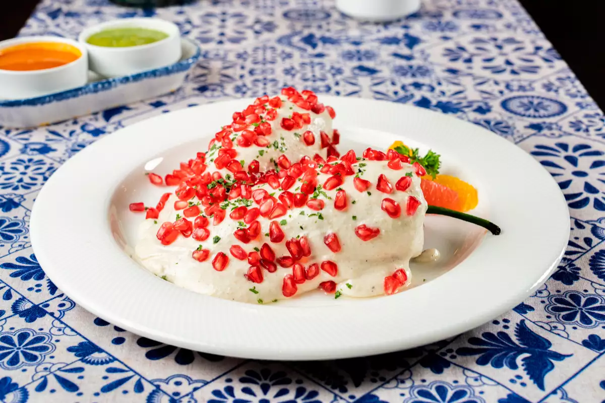 Foto chile en nogada servido en un plato blanco extendido sobre una mesa con mosaicos de talavera azul y blanca.