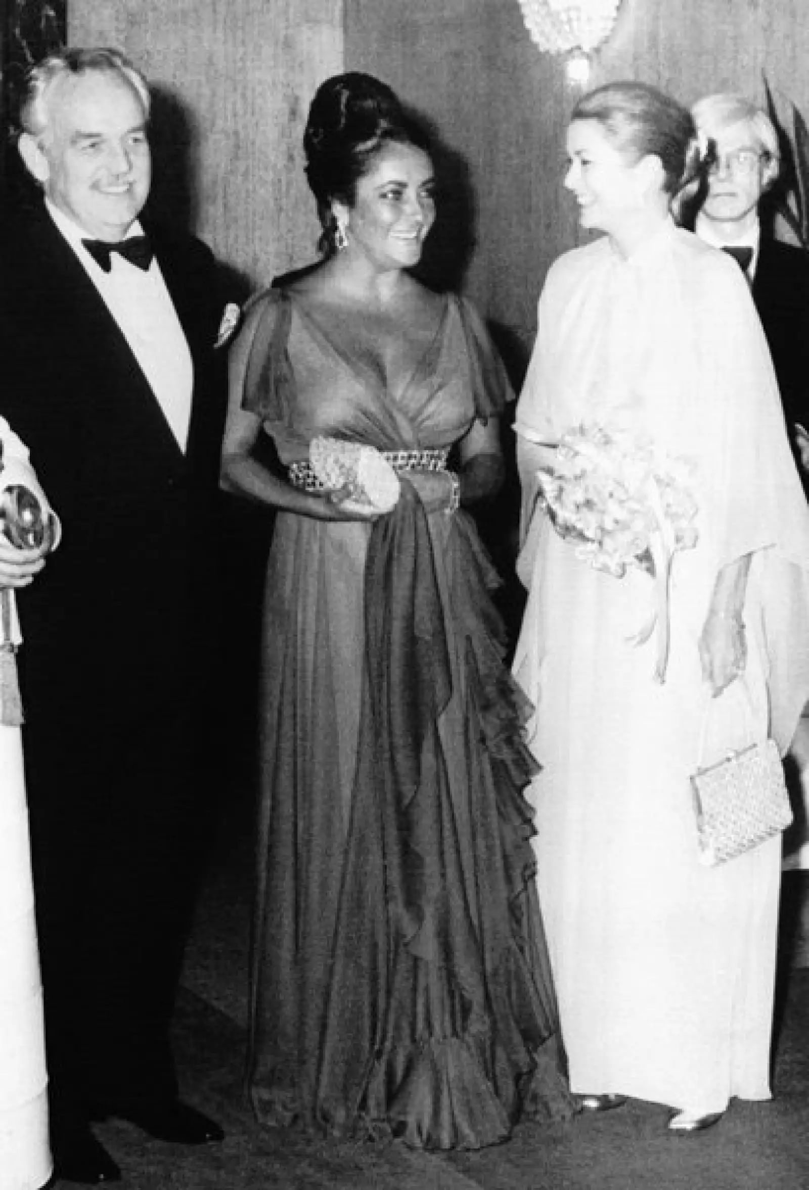 Elizabeth Taylor platica animadamente con los príncipes de Mónaco durante un evento de beneficiencia de la Cruz Roja el 21 de mayo de 1974. El artista y cineasta Andy Warhol está detrás de Grace Kelly.