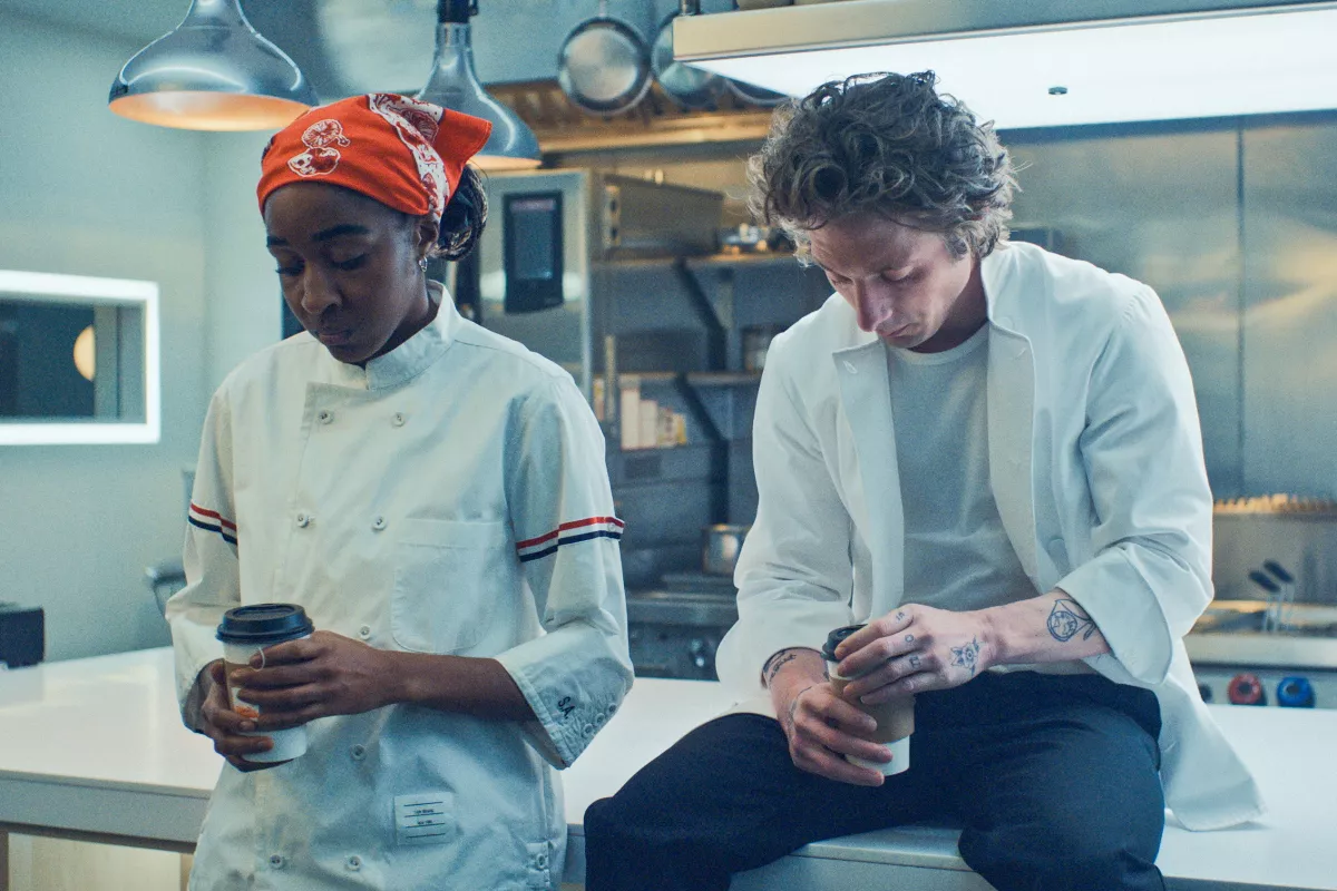 Foto de la serie The Bear con Sydney (izquierda) y Carmy (derecha) sentados sobre una plancha de cocina y con tazas de café entre sus manos.