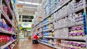 Interior de un supermercado con un amplio pasillo dedicado a productos de papel y artículos de limpieza. A ambos lados del pasillo se observan estanterías metálicas altas, completamente abastecidas con paquetes de papel higiénico, servilletas, toallas de papel y productos de limpieza de distintas marcas. En el lado derecho predominan grandes cantidades de rollos y paquetes de papel higiénico, mientras que en el lado izquierdo se exhiben productos de limpieza y paquetes de artículos para el hogar. En el centro del pasillo, un trabajador del supermercado, vestido con pantalón de mezclilla, camiseta blanca y chaleco rojo, se encuentra agachado frente a los estantes mientras acomoda mercancía. 