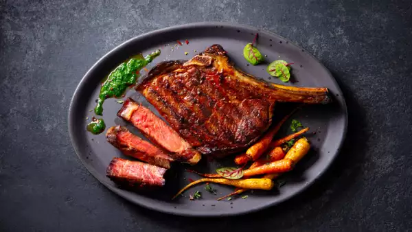Beef tomahawk steak on a bone with roasted colorful carrots and chimichurri sauce on a plate. Grey background. Close up. Top view.