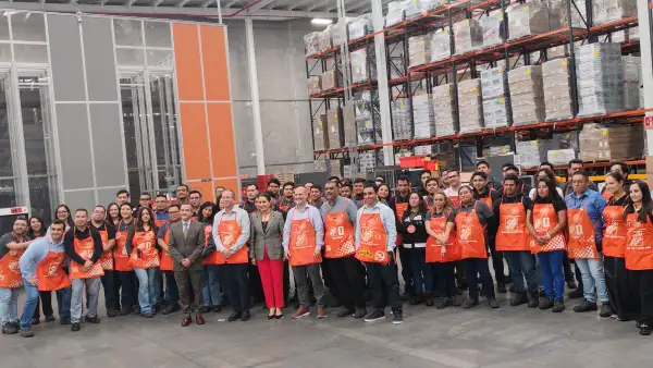 Personal de Home Depot vistiendo los mandiles de dependientes de la cadena en color naranja con el logo de la cadena en color blanco. Están en uno de los centros de distribución de la empresa por lo que hay algunos racks en la parte del fondo. 