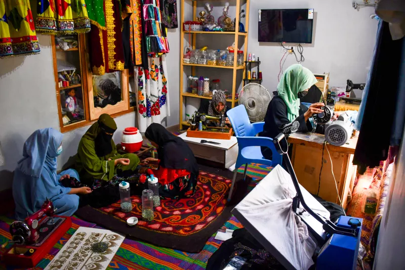 Mujeres afganas en un taller de confección de ropa, es una habitación bastante pequeña y hay 5 mujeres cubiertas por burkas. De izquiera a derecha, lleva una burka azul, la segunda lleva una burka verde, la tercera burka rojo con negro, la cuarta casi no se ve pues esta detrás de una maquina de coser y la quinta, que esta sentada en una silla, lleva negro con verde menta.