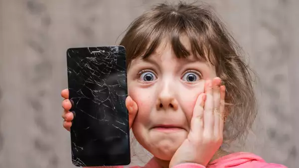 The frightened child accidentally ruined the smartphone. Portrait of a scared little girl with a broken mobile phone. A sad child broke the screen of a mobile phone. Cracked display in hand kids.