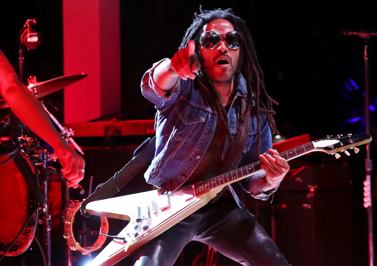 Lenny Kravitz usando lentes oscuros y señalando al frente con el dedo índice mientras con la otra mano sostiene su guitarra.