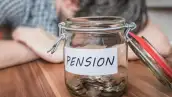 Persona recostada sobre una mesa y un tarro de vidrio con monedas y el emblema pensión.