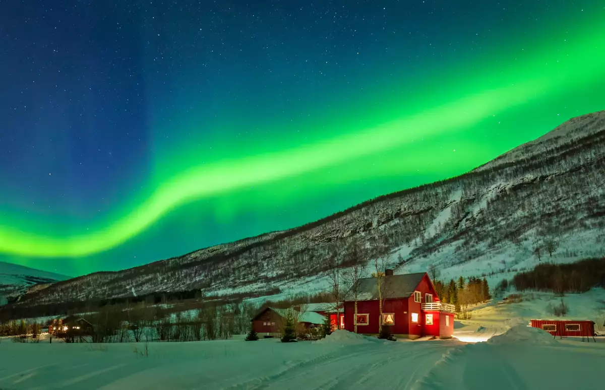 Foto de una casa en un pueblo rural de Noruega y se observa una aurora boreal verde en el cielo.
