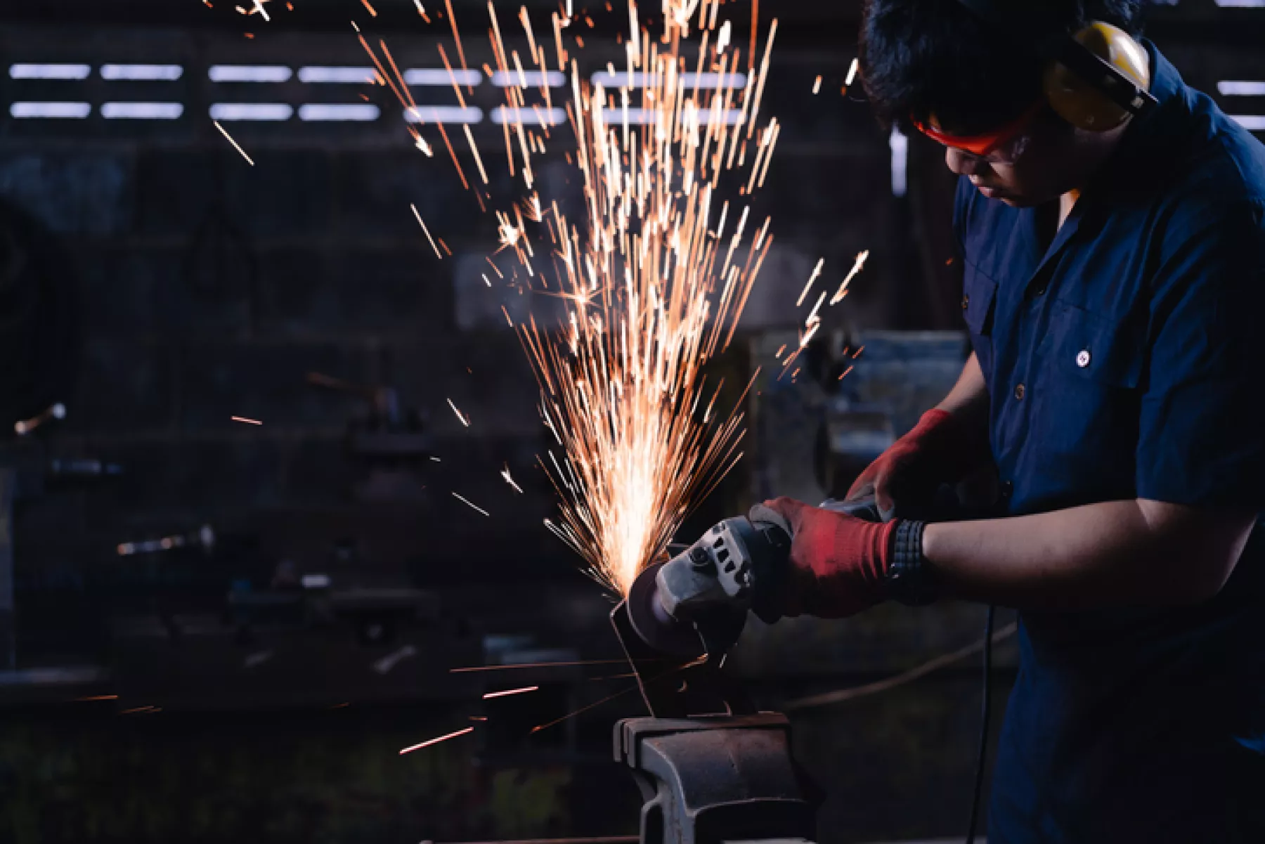 Immigrant manufacturing worker using angle grinder power tools on metal creating hot sparks wearing safety protective work wear