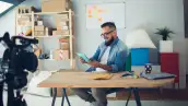 Un hombre con barba y lentes sentado frente a una mesa viendo una libreta, en primer plano una cámara de video.jpg