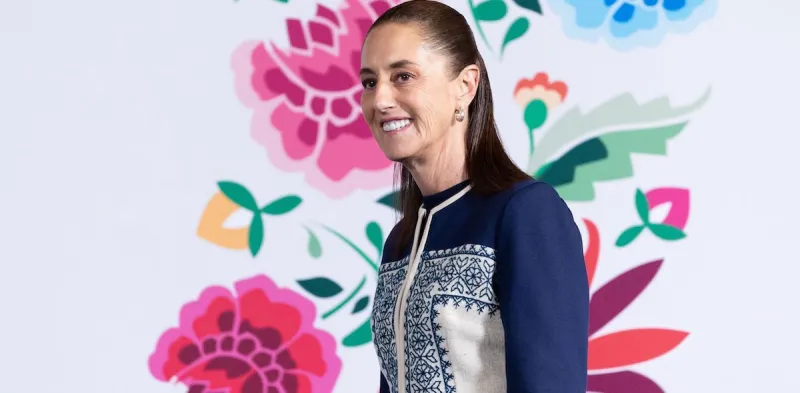 La presidenta Claudia Sheinbaum sonríe a los asistentes de la conferencia mañanera en Palacio Nacional. Hay un fondo blanco con rosas pintadas. 