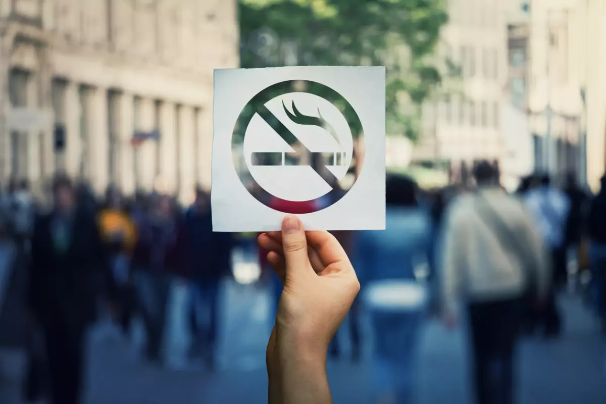 Mano sosteniendo una hoja de papel con un cartel de no fumar sobre un fondo de lugar público en una calle concurrida. Zona de área prohibida, símbolo restrictivo detener el humo.