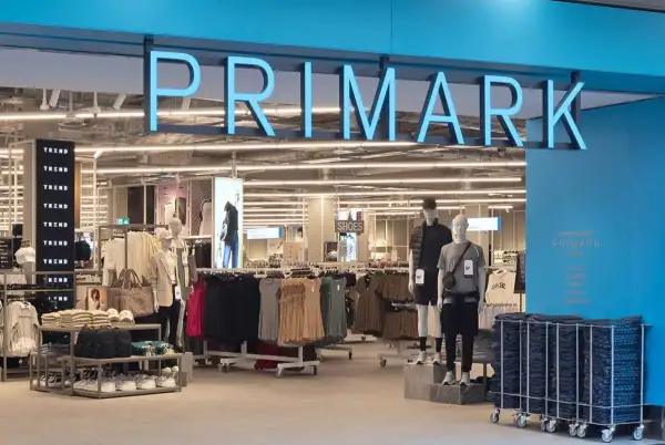 Entrada de una tienda Primark, en la que se muestran aparadores con ropa y zapatos de colores neutros como gris y nude. La fachada es color azul, con el nombre de la cadena en el mismo color en la parte de arriba de la entrada.

