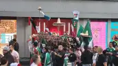 En la imagen se ve un carrito de venta de banderas, mientras personas caminan frente a este. 