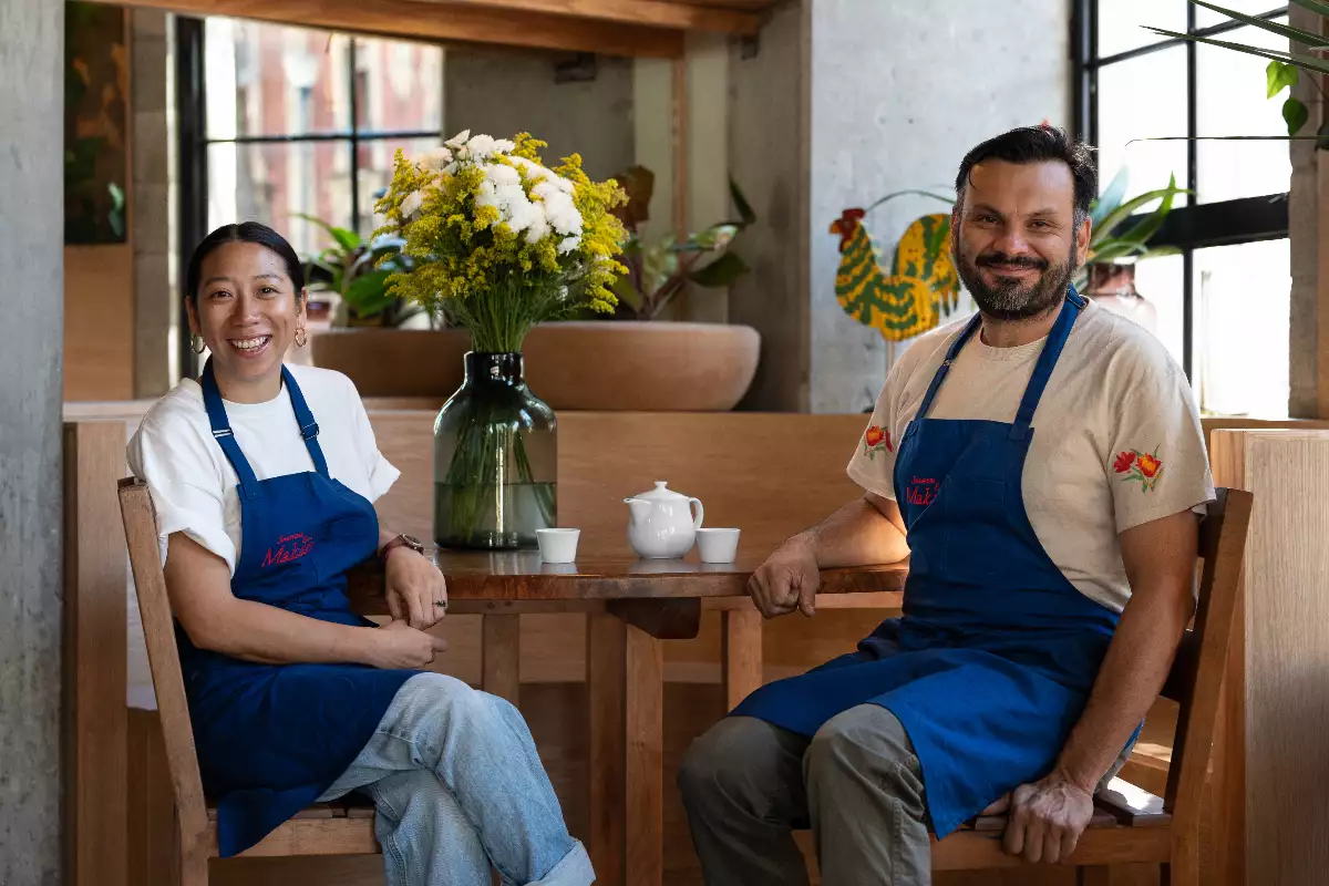 Foto de los chefs Maryann Yong y Mario Malváez sentados en su restaurante Makan, sonriendo a la cámara y vistiendo playeras blancas y mandiles azules.