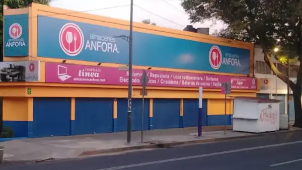 Sucursal de Almacenes Ánfora sobre una avenida, con una fachada de dos niveles en azul y naranja, el logotipo de la empresa visible en distintos puntos y anuncios de categorías como repostería, loza restaurantera, sartenes, electrodomésticos, cristalería, baterías de cocina y vajillas. La entrada tiene cortinas metálicas cerradas y al frente se observan una banqueta, postes, árboles y un pequeño puesto, en una escena urbana.