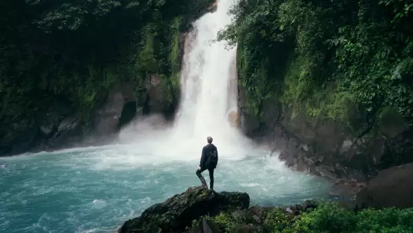 Foto de un hombre parado sobre unas rocas frente a una cascada en el bosque.