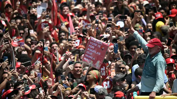 El presidente Luiz Inácio Lula da Silva saluda a una multitud de simpatizantes durante un acto de campaña en Brasil. Los asistentes, vestidos principalmente de rojo, levantan sus teléfonos para fotografiarlo y portan banderas y carteles con el número 13, asociado a su candidatura. Lula viste una camisa azul claro y una gorra roja mientras avanza entre la concentración.