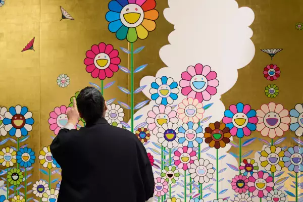 Foto de Takashi Murakami de espalda a la cámara observando un muro intervenido con sus icónicas flores sonrientes.
