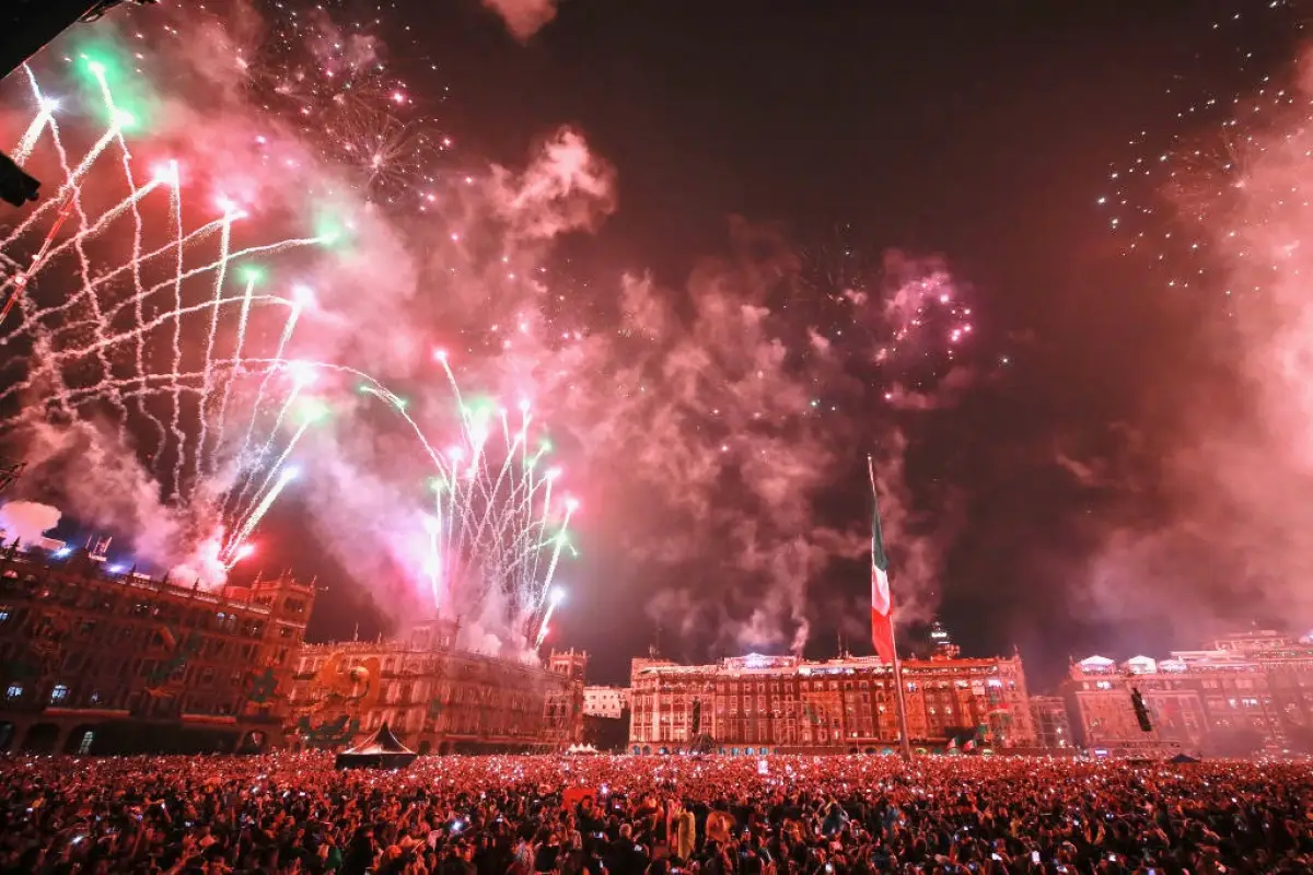 Fuegos artificiales en verde, blancoo y rojo sobre el zócalo de la CDMX con la bandera de México al fondo durante el grito de independencia.