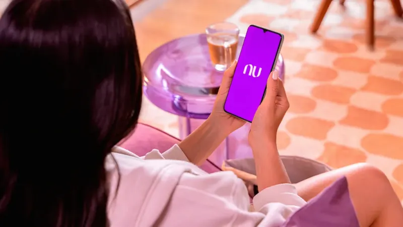 Mujer de espaldas, mientras sostiene con ambas manos su celular en la que se ve su celular con la pantalla de inicio de la aplicación de Nu.