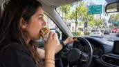 Mujer conduce un automóvil en una avenida de la Ciudad de México mientras da una mordida a una torta y mantiene una mano sobre el volante.