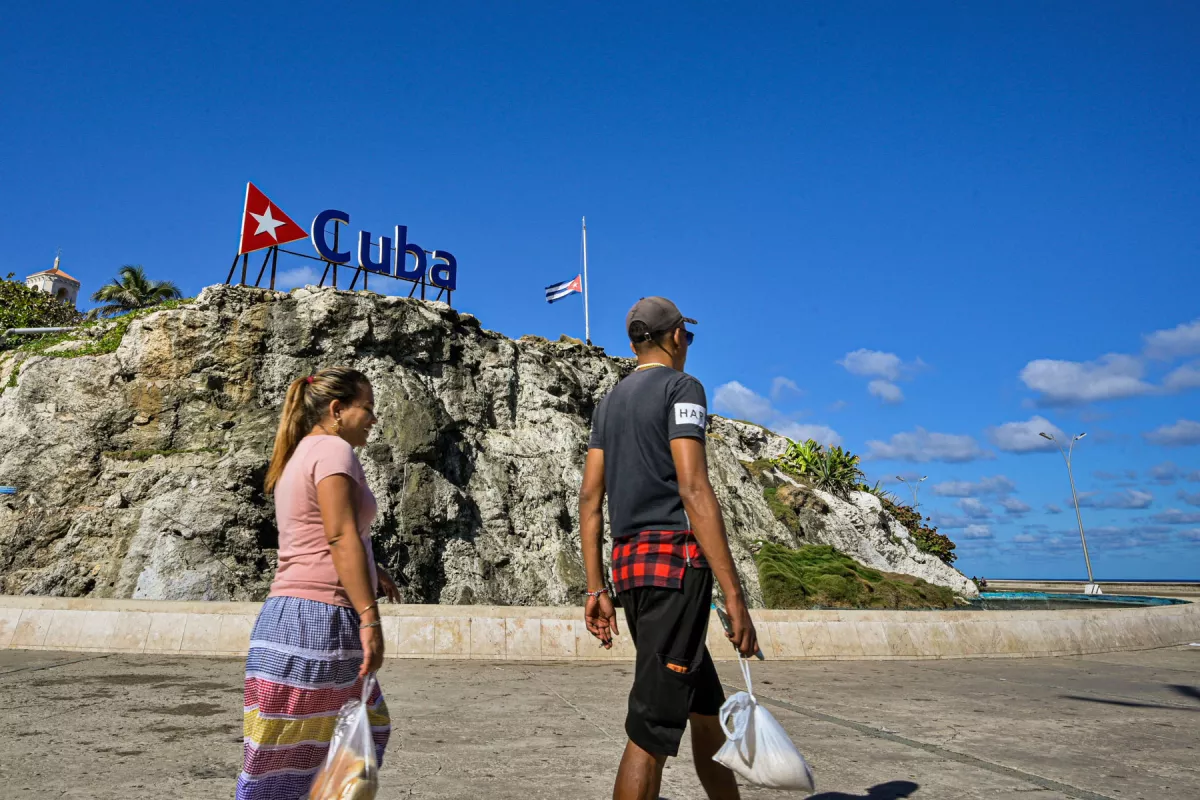 Los cubanos caminan por un cartel de Cuba y una bandera nacional a media asta en La Habana el 5 de enero de 2026. La Habana declaró dos días de luto nacional a partir del 5 de enero después de que un total de 32 cubanos fueran asesinados durante el ataque estadounidense a Caracas que culminó con la captura del presidente venezolano Nicolás Maduro.