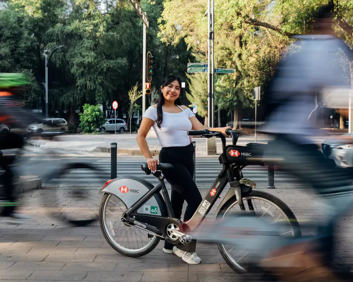 Una joven tomando una Ecobici posa para la foto mientras en el fondo se ve la silueta barrida de otros ciclistas que pasan.    