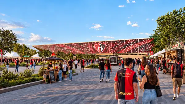 Render exterior del nuevo estadio del Atlas, con aficionados caminando por una explanada rodeada de áreas verdes, comercios y puestos de comida.