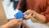 El brazo de una mujer sosteniendo una tarjeta de crédito para pagar una compra, junto con la mano de otra persona que acepta el pago en una terminal bancaria