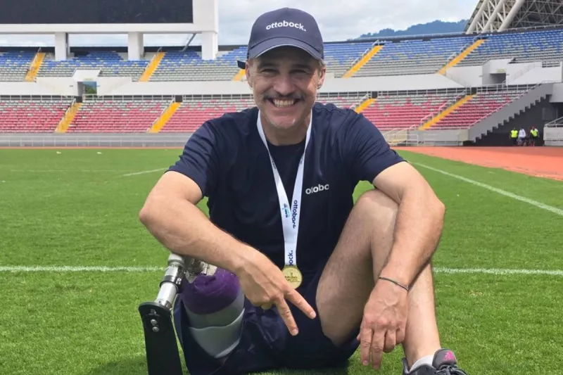 Foto del actor Juan Pablo Medina sonriendo y sentado en un campo de futbol, usa una gorra y muestra su prótesis deportiva.