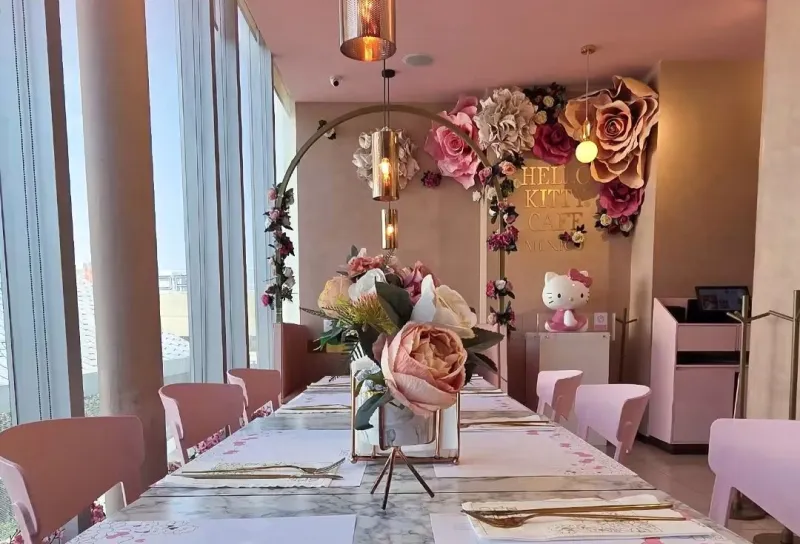 Una mesa con manteles blancos y un arreglo de rosas de diferentes tonalidades de rosa. Al fondo una Hello Kitty y arriba un letrero en color dorado con las palabras Hello Kitty Café.