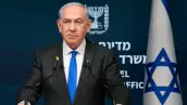 Benjamin Netanyahu, un hombre blanco de 74 años. Cabello blanco canoso, viste un traje negro, una camisa blanca y corbata azul vibrante. Porta una expresión seria. Detrás de él, una bandera de Israel.