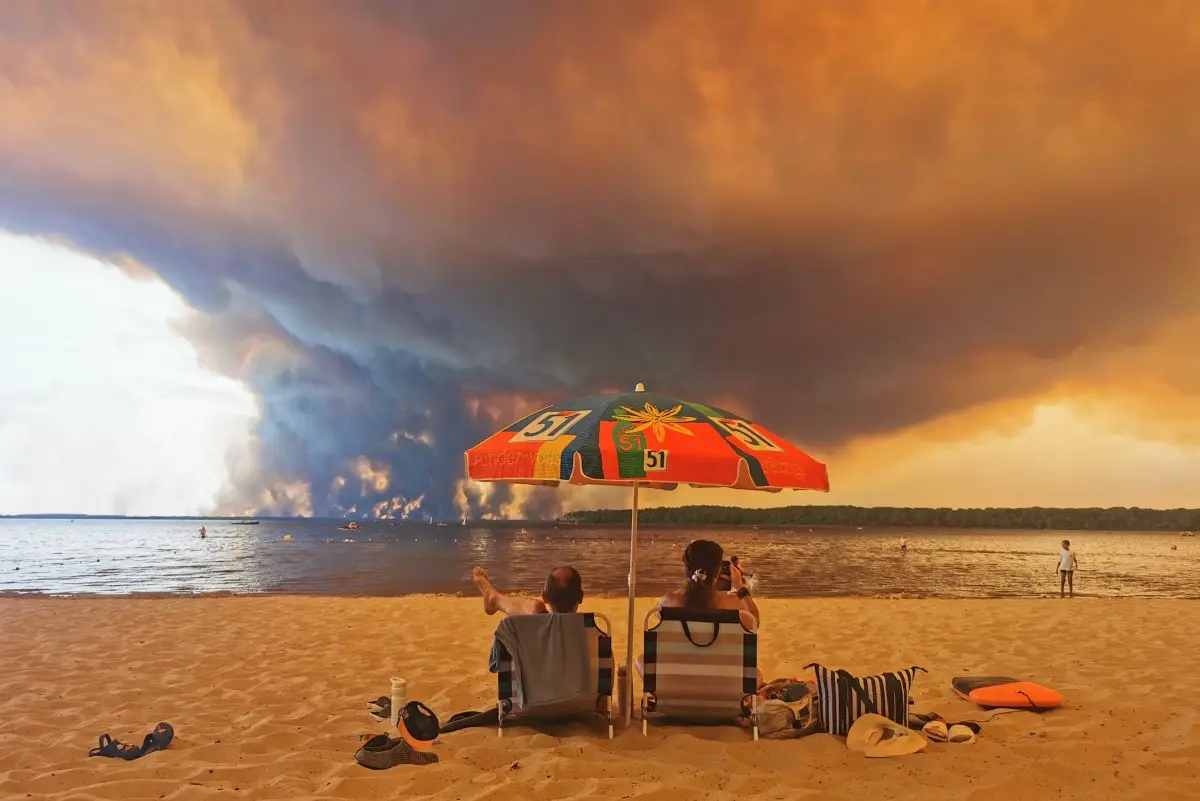 Los bañistas se sientan en la playa de Moutchic en Lacanau, al suroeste de Francia, el 24 de julio de 2026, mientras las nubes de humo se elevan al cielo a partir de un incendio forestal.
