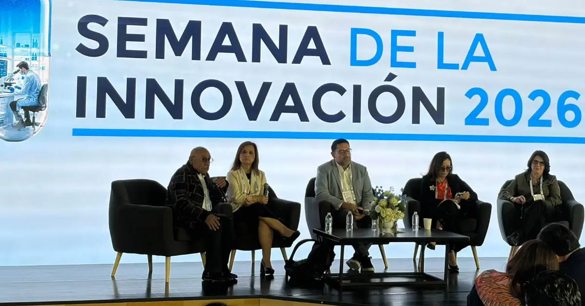 Dos hombres de traje y tres mujeres con vestidos formales frente una mesa de centro. Un letrero con Semana de la Innovación.jpg