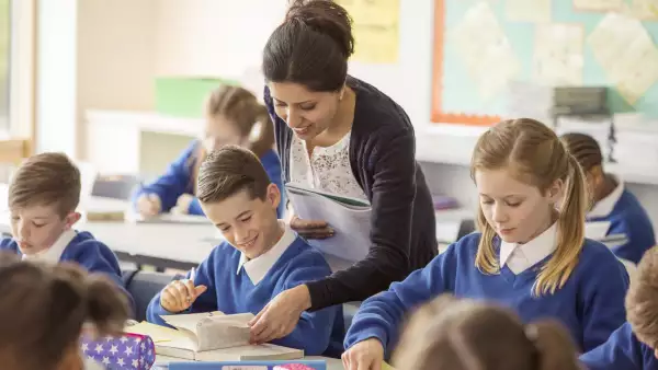 Profesora de primaria muestra una página de un libro a un niño vestido con un suéter azul. 