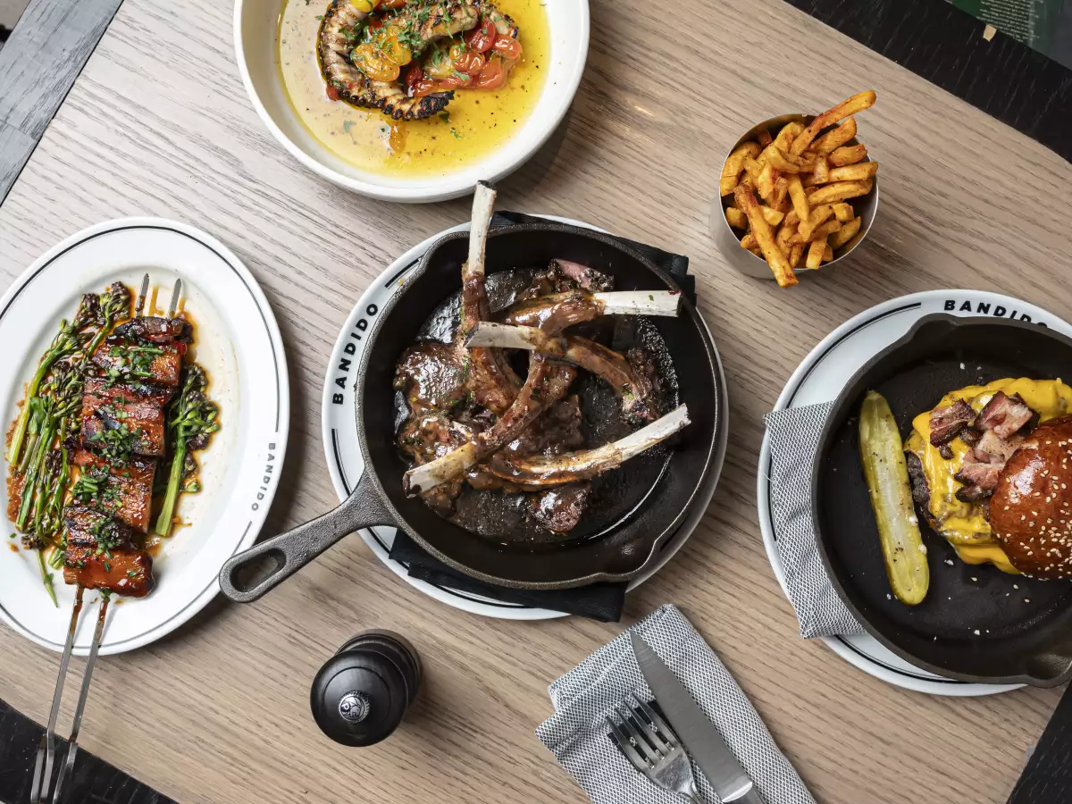 Foto de una mesa con comida del restaurante Bandido, al centro unas costillas, a los lados una hamburguesa, papas a la francesa y un plato con pulpo a las brasas y jitomates cherry.