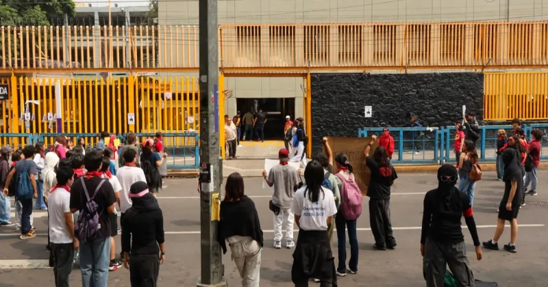 Grupo de jóvenes de espaldas frente a unas rejas amarillas de un plantel de la UNAM.jpg