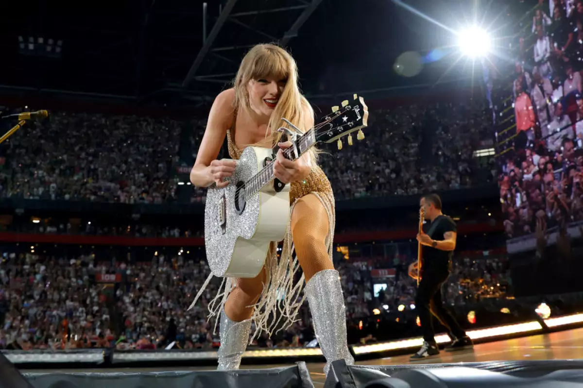 Foto de Taylor Swift sonriendo agachada hacia el frente con una guitarra acústica en sus manos durante un concierto en Amsterdam.