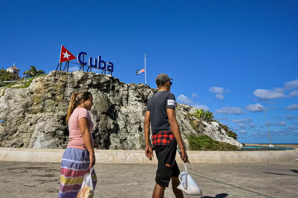Los cubanos caminan por un cartel de Cuba y una bandera nacional a media asta en La Habana el 5 de enero de 2026. La Habana declaró dos días de luto nacional a partir del 5 de enero después de que un total de 32 cubanos fueran asesinados durante el ataque estadounidense a Caracas que culminó con la captura del presidente venezolano Nicolás Maduro.