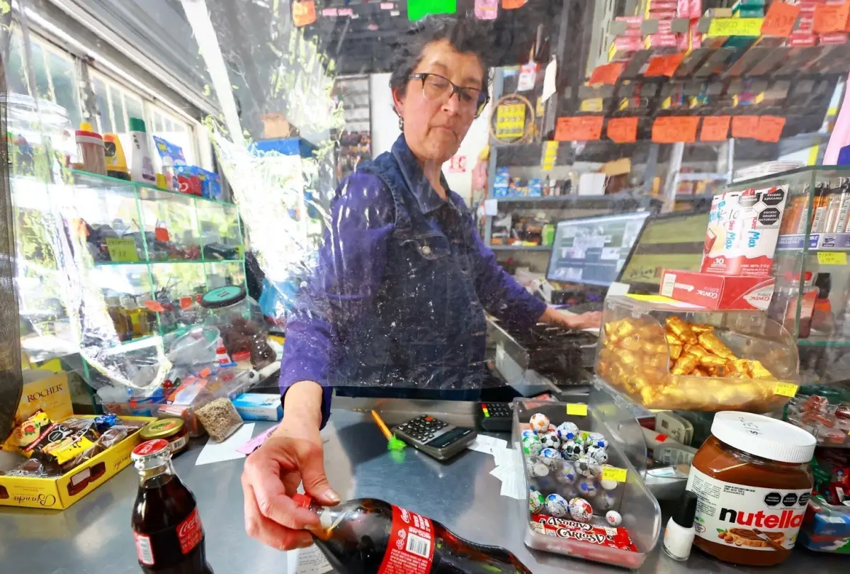 Una mujer de cabello corto y oscuro vestida con una blusa morada detrás de un mostrador cobra refrescos a un cliente.