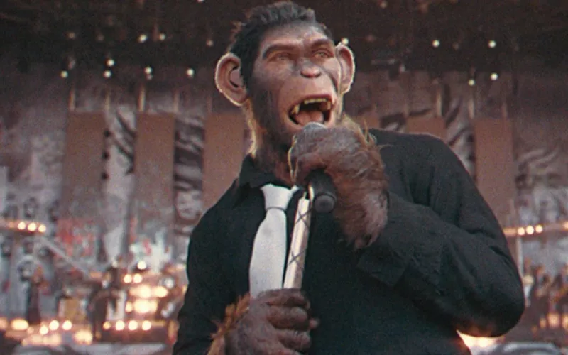 Foto de una escena de Better Man, con un simio vestido de camisa negra y corbata blanca y un micrófono en la mano.
