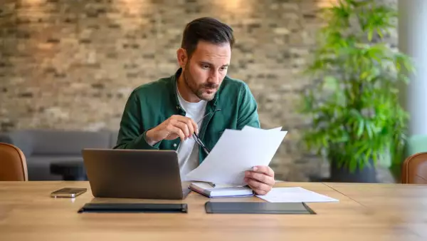 Serious male accountant holding glasses and analyzing financial report with laptop on desk in office