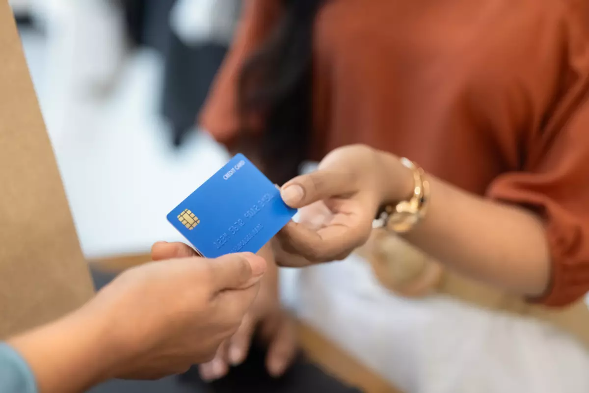 El brazo de una mujer sosteniendo una tarjeta de crédito para pagar una compra, junto con la mano de otra persona que acepta el pago en una terminal bancaria