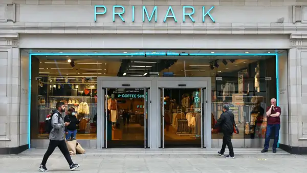 Entrada de una tienda Primark, en la que se muestran aparadores con ropa y zapatos de colores neutros como gris y nude. La fachada es color azul, con el nombre de la cadena en el mismo color en la parte de arriba de la entrada.

