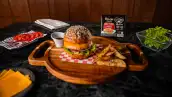 Un a hamburguesa de salmón con papás fritas sobre un papel encerado de cuadros color blanco y rojo, que está encima de una tabla redonda de madera. Del lado superior derecho hay un paquete de las carnes para la hamburguesa de la marca Dolores, junto hay servilletas, un plato color negro con jitomate rebanado, otro con queso amarillo y un refractario de cristal con lechuga. 