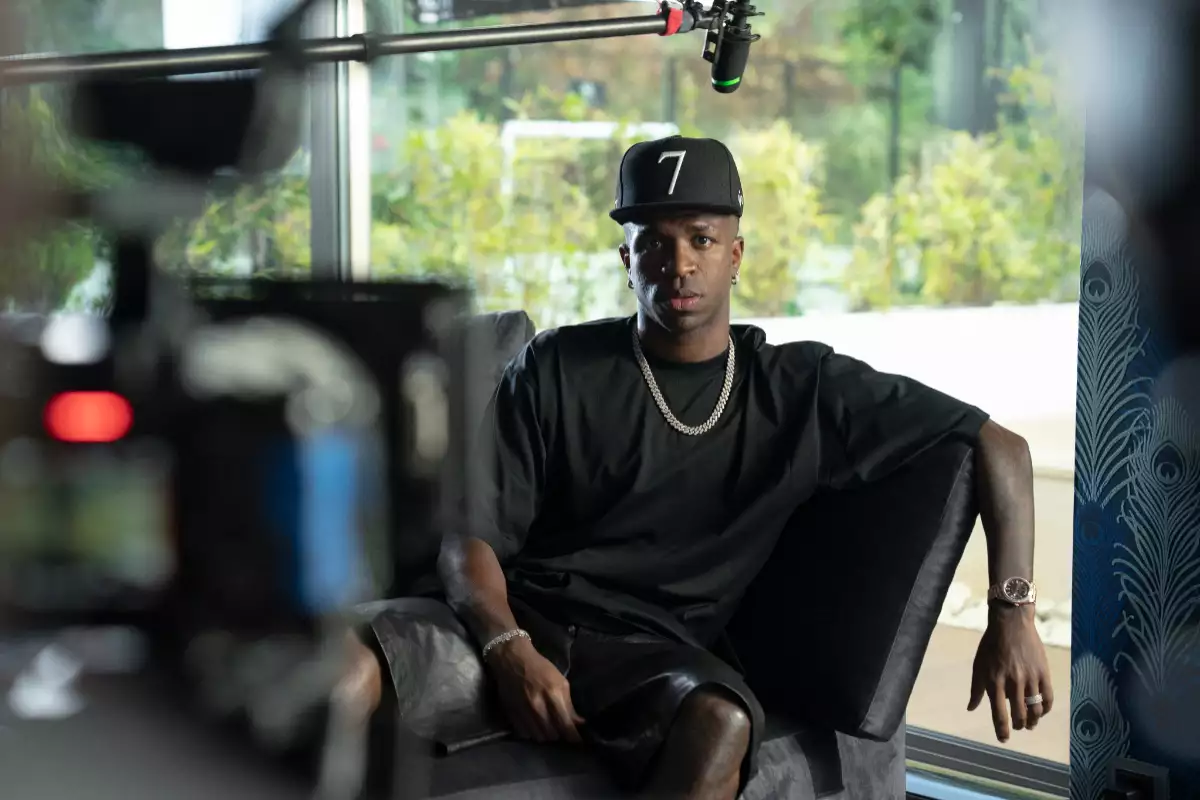 Foto de Vinicius Jr vestido casual de negro con gorra y frente a la cámara de cine que graba su documental.