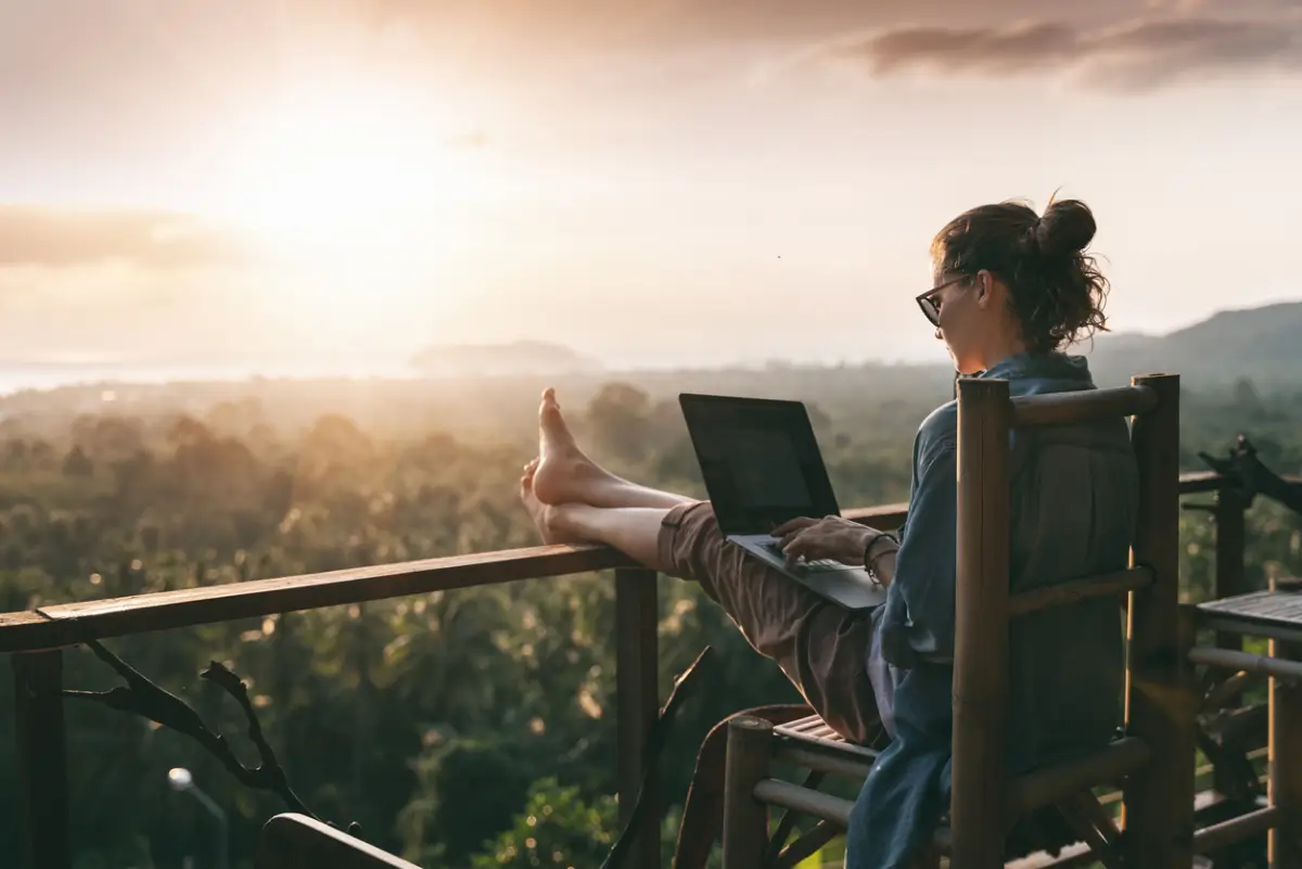 Las visas para nómadas digitales ya son una realidad en estos países. (Mujer sentada con las piernas estiradas y una laptop sobre ellas, con vista a un bosque). 