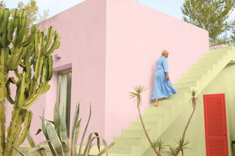 Una mujer asciende por unas escaleras en un edificio pintado con color plasma, un tono rosado. Al frente se observan unas plantas.