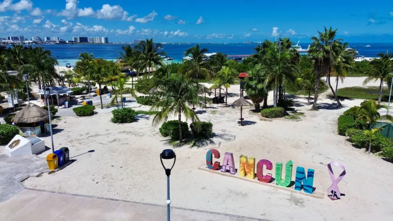 Vista panorámica de una zona costera de Cancún, con un letrero multicolor que forma el nombre de la ciudad y un listón morado a un costado. Al fondo se observan palmeras, arena blanca, embarcaciones y el mar Caribe, bajo un cielo azul despejado.