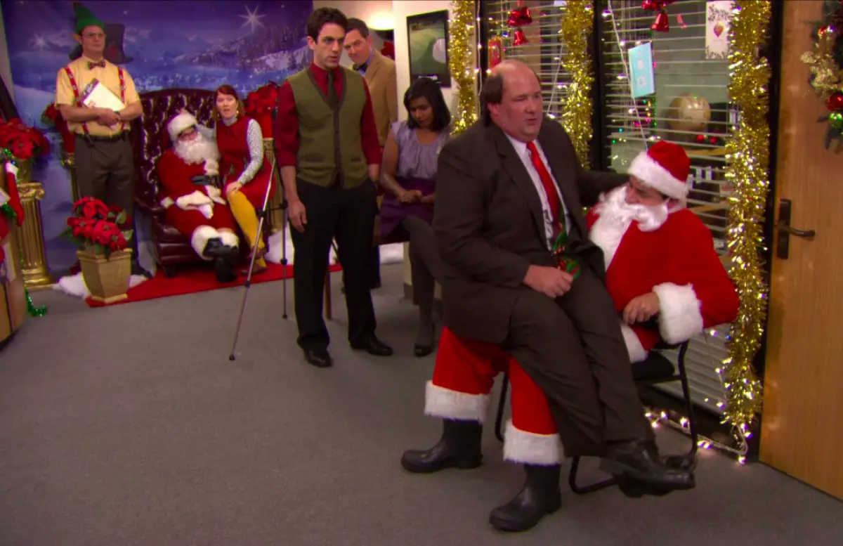 Foto de una escena de The Office con Michael Scott vestido de Santa Claus y Kevin sentado en sus piernas